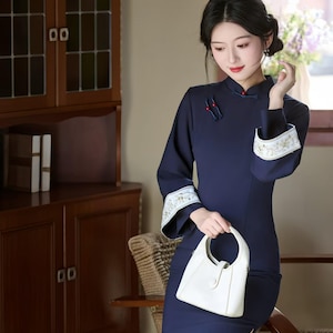 May include: Navy blue, long-sleeve dress with a mandarin collar and white embroidered cuffs. The dress features a side slit and is paired with a white handbag. The woman is sitting in a chair in front of a wooden cabinet.
