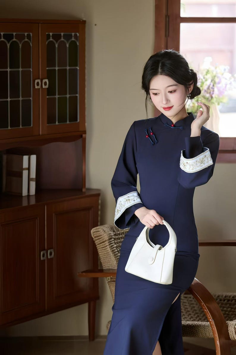 May include: Navy blue, long-sleeve dress with a mandarin collar and white embroidered cuffs. The dress features a side slit and is paired with a white handbag. The woman is sitting in a chair in front of a wooden cabinet.