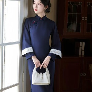 May include: A navy blue, long-sleeved dress with a mandarin collar and white floral embroidery on the cuffs. The dress is paired with a white handbag. The woman is standing near a window.