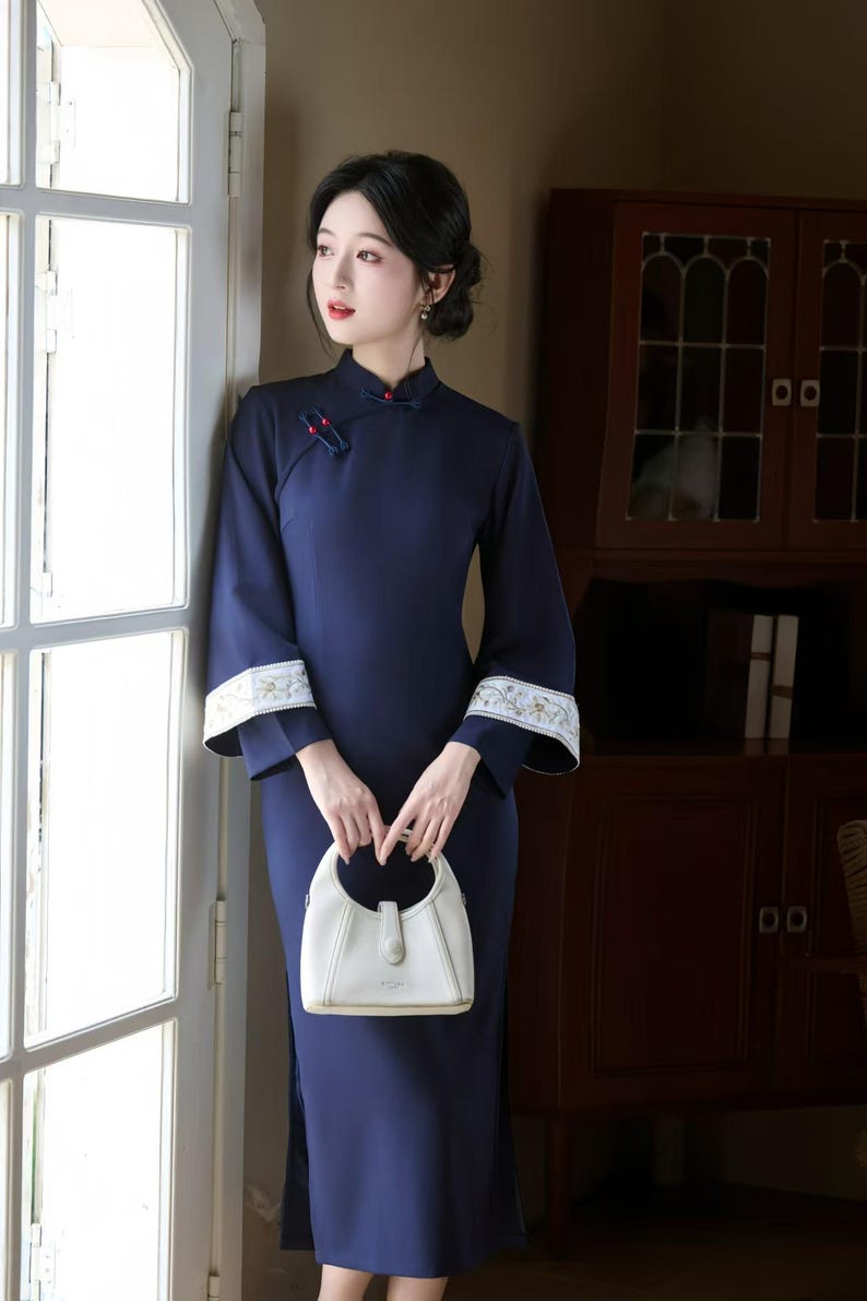 May include: A navy blue, long-sleeved dress with a mandarin collar and white floral embroidery on the cuffs. The dress is paired with a white handbag. The woman is standing near a window.