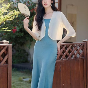 May include: A woman wearing a light blue maxi dress and a cream-colored bolero jacket. She is holding a fan. The background includes a wooden fence and greenery.