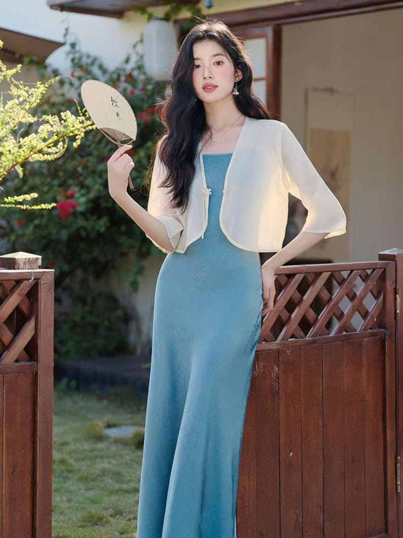 May include: A woman wearing a light blue maxi dress and a cream-colored bolero jacket. She is holding a fan. The background includes a wooden fence and greenery.