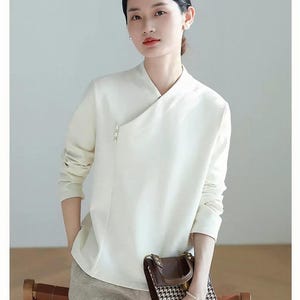 Pode incluir: Uma blusa branca de manga comprida com gola mandarim e um detalhe de bot&atilde;o de p&eacute;rola &uacute;nico. A blusa tem um caimento ligeiramente solto e est&aacute; enfiada em cal&ccedil;as bege.