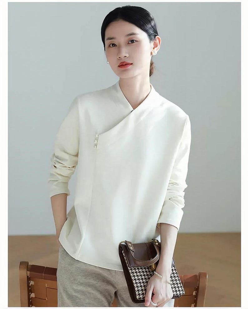 Pode incluir: Uma blusa branca de manga comprida com gola mandarim e um detalhe de bot&atilde;o de p&eacute;rola &uacute;nico. A blusa tem um caimento ligeiramente solto e est&aacute; enfiada em cal&ccedil;as bege.
