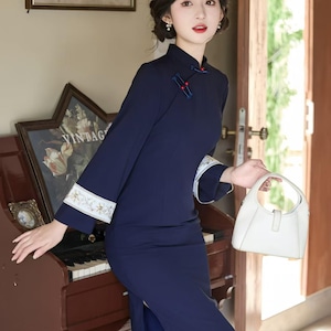 May include: A navy blue, long-sleeved dress with a mandarin collar and side slit. The dress features white embroidered cuffs and red accents. A white handbag is held in the hand. The dress is a vintage style.