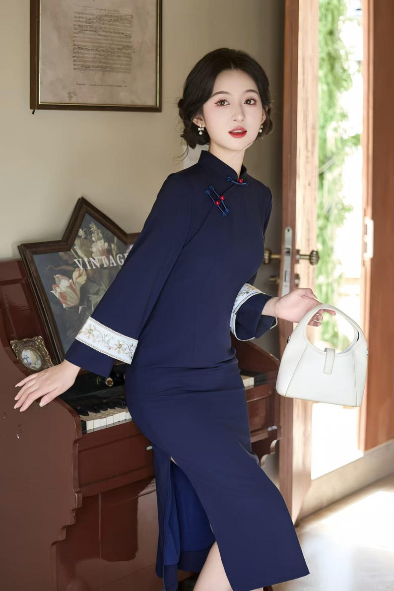 May include: A navy blue, long-sleeved dress with a mandarin collar and side slit. The dress features white embroidered cuffs and red accents. A white handbag is held in the hand. The dress is a vintage style.