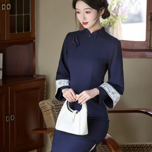 May include: A navy blue, long-sleeved dress with a mandarin collar and a side slit. The dress features embroidered cuffs and is paired with a white handbag. The style is reminiscent of traditional Chinese attire.