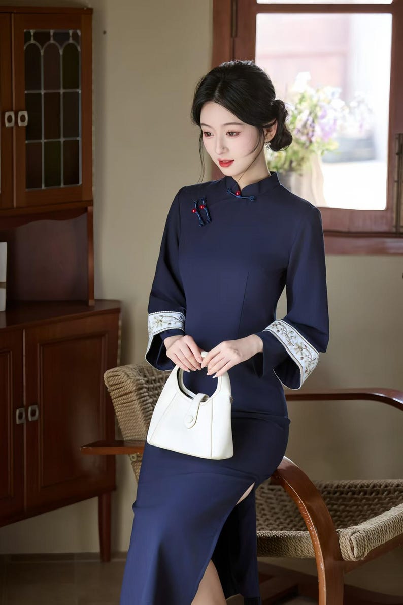 May include: A navy blue, long-sleeved dress with a mandarin collar and a side slit. The dress features embroidered cuffs and is paired with a white handbag. The style is reminiscent of traditional Chinese attire.