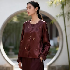 May include: A burgundy silk blouse and matching pants. The blouse has long sleeves, a round neckline, and small gold embroidered details. The outfit is worn by a person with dark hair pulled back in a ponytail.