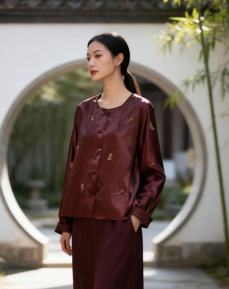 May include: A burgundy silk blouse and matching pants. The blouse has long sleeves, a round neckline, and small gold embroidered details. The outfit is worn by a person with dark hair pulled back in a ponytail.