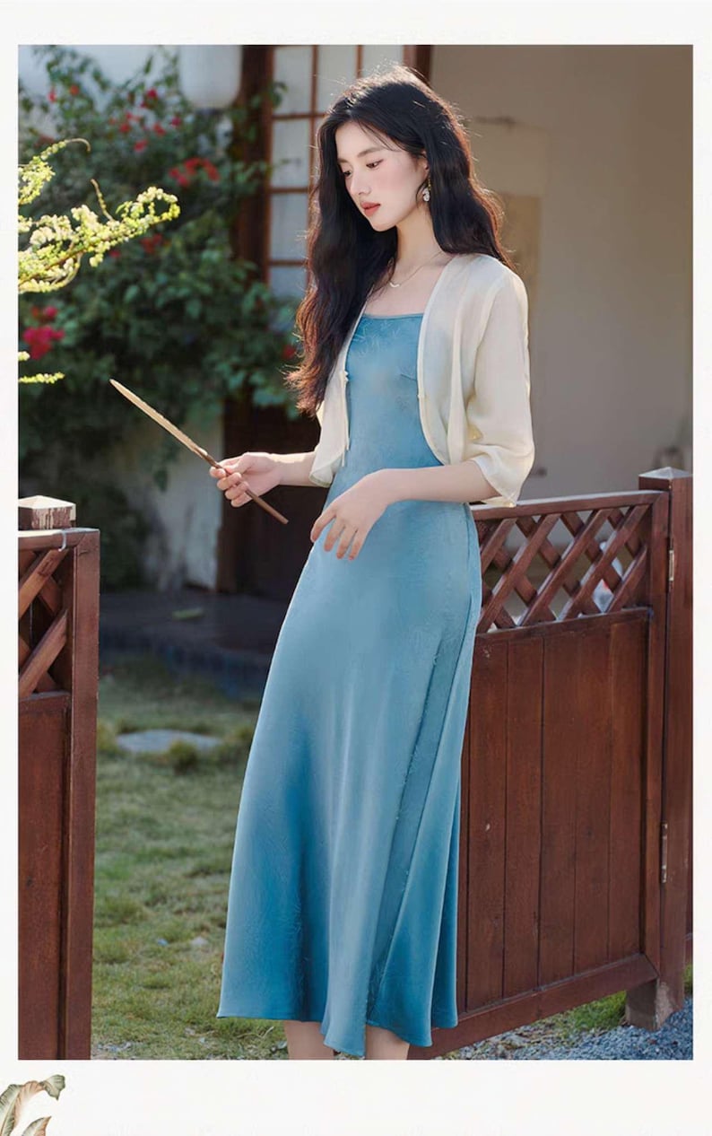 May include: A long, flowing, light blue dress with a square neckline and a side slit. A short, cream-colored bolero jacket is worn over the dress. The outfit is completed with delicate jewelry. The background features a wooden fence and greenery.