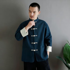 Può includere: Giacca blu scuro in stile Tang con chiusure e polsini bianchi. La giacca ha un colletto alla coreana e due tasche anteriori. Le maniche sono a tre quarti. La giacca è indossata sopra una camicia nera.