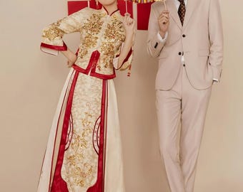 May include: A couple in traditional Chinese wedding attire. The woman wears a cream-colored dress with gold embroidery and red accents. The man wears a light beige suit with a patterned tie. Both hold decorative items. The background features a red double happiness symbol.