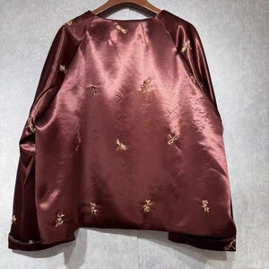 May include: A maroon satin jacket with gold embroidered floral designs. The jacket has long sleeves and a round neckline. The cuffs and hem are trimmed with a darker maroon velvet. The jacket is hanging on a wooden hanger.