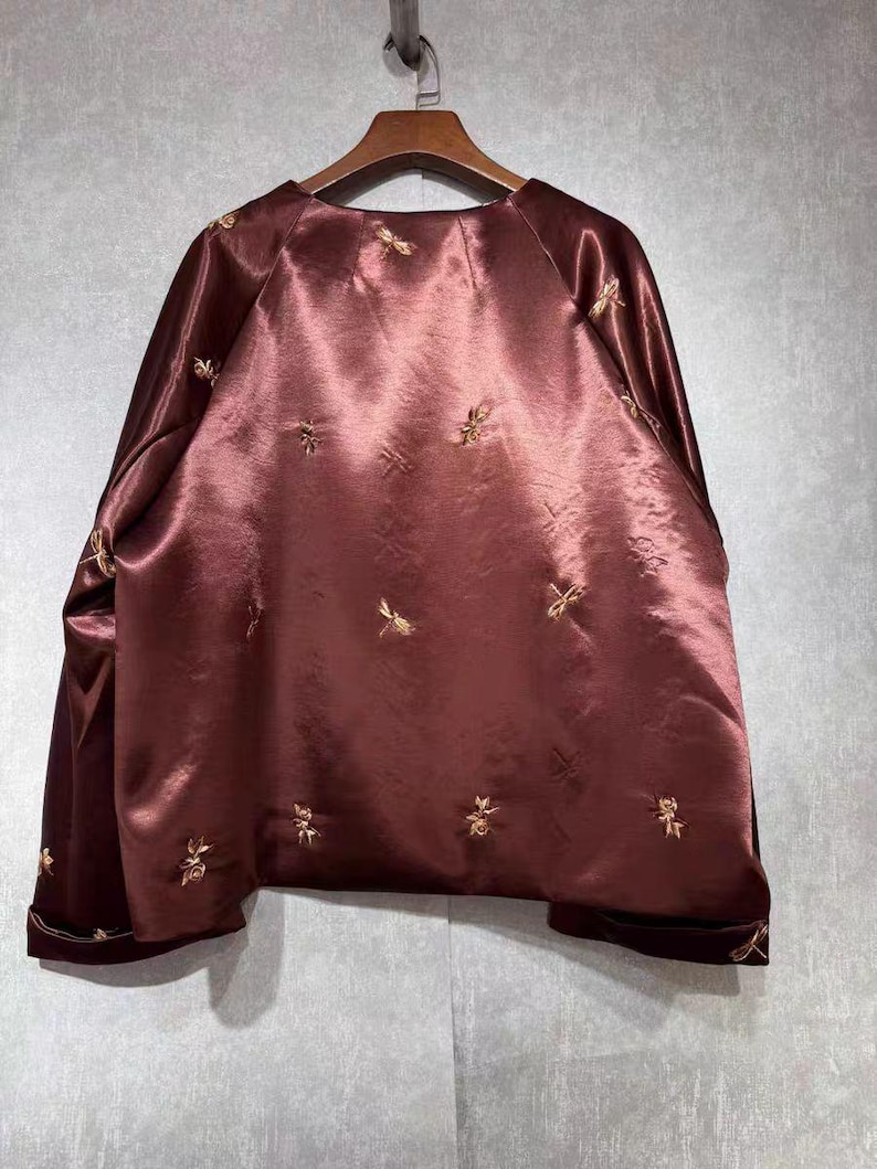 May include: A maroon satin jacket with gold embroidered floral designs. The jacket has long sleeves and a round neckline. The cuffs and hem are trimmed with a darker maroon velvet. The jacket is hanging on a wooden hanger.