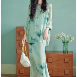 Puede incluir: Un conjunto de dos piezas con una camisa de manga larga con botones y una falda a juego, ambas con un estampado floral de acuarela en verde claro y azul. El conjunto se combina con un bolso blanco y tacones verdes. Un look elegante y con estilo.