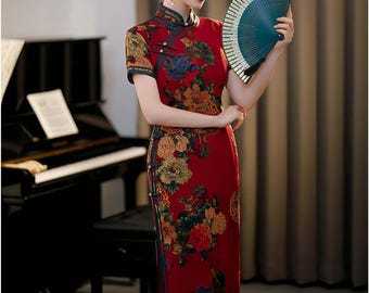 May include: A red cheongsam dress with a floral print and a high collar. The dress has short sleeves and a side slit. The model is holding a blue and white hand fan.