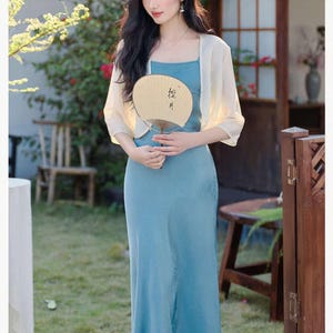 May include: A woman wearing a light blue spaghetti strap dress and a cream-colored shawl, holding a beige fan. The dress is floor-length. The image has a soft, natural lighting and a blurred background.