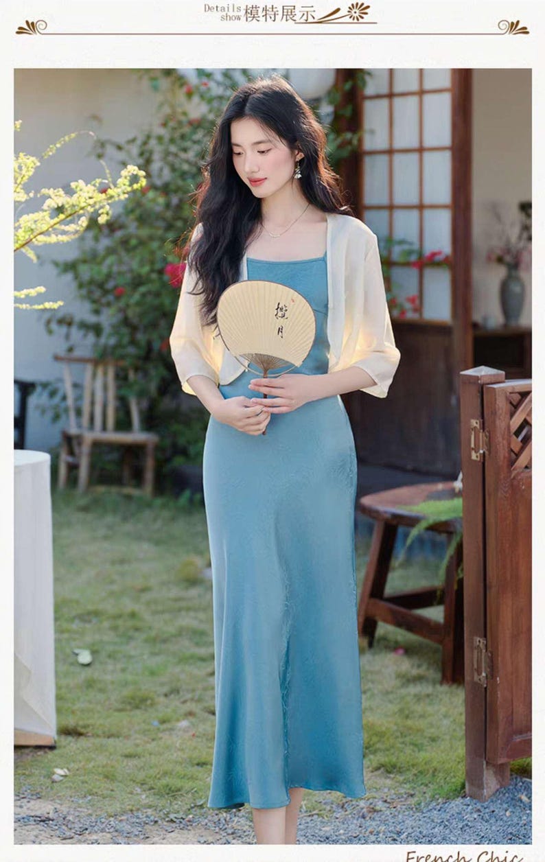 May include: A woman wearing a light blue spaghetti strap dress and a cream-colored shawl, holding a beige fan. The dress is floor-length. The image has a soft, natural lighting and a blurred background.