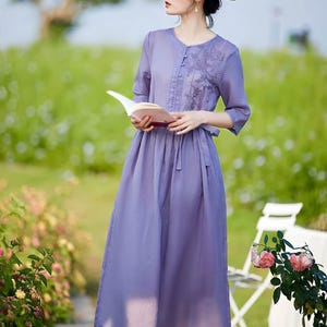 Peut inclure: Une robe lavande avec un col rond et des manches trois-quarts. La robe présente des détails brodés sur le corsage et une jupe fluide. Un chapeau de paille et un livre sont également visibles. La robe est adaptée aux tenues décontractées.