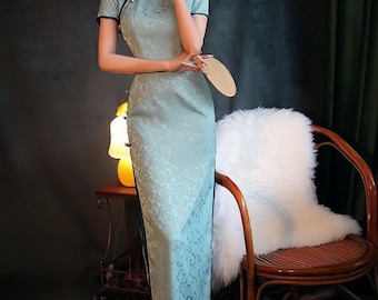 May include: A light blue, short-sleeved, traditional Chinese dress with a floral pattern. The dress has a mandarin collar and frog closures. The model is holding a small fan. The dress is floor-length and form-fitting.
