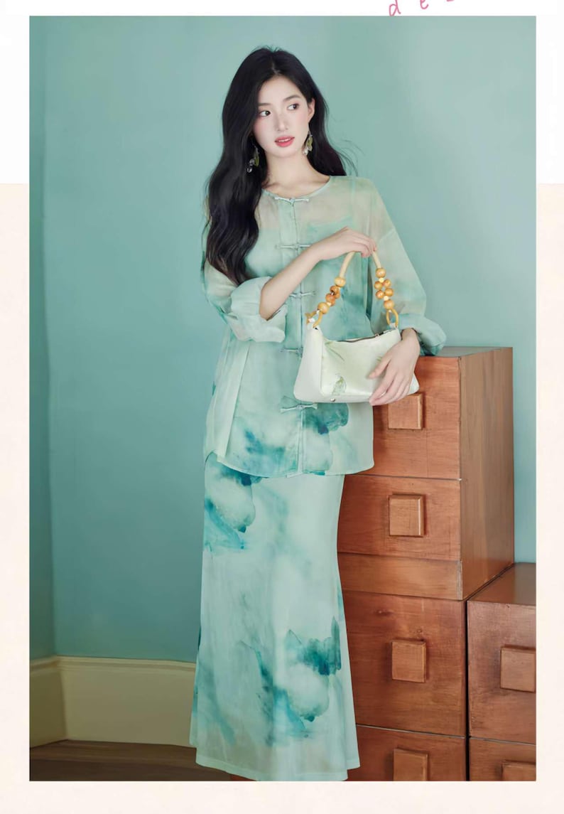 Puede incluir: Un conjunto de dos piezas verde menta con un dise&ntilde;o de acuarela. El conjunto incluye una blusa de manga larga con cierres de rana y una falda a juego. Se sostiene un bolso de mano blanco con asa de bamb&uacute;.