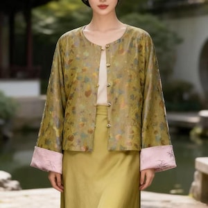 Puede incluir: Una mujer con una chaqueta verde con estampado floral, una blusa crema y una falda verde. La chaqueta tiene puños rosas con detalles bordados. El atuendo es una mezcla de estilos tradicionales y modernos.