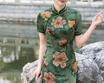 May include: A green, short-sleeved dress with a floral pattern featuring large, tan and yellow flowers. The dress has a mandarin collar and frog closures. The model is holding a black fan. The dress is a traditional Chinese garment.