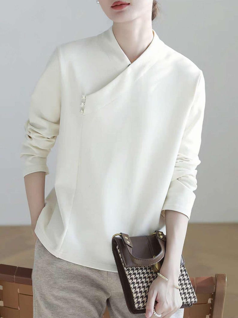 Pode incluir: Uma blusa branca de manga comprida com gola mandarim e fecho diagonal. A blusa tem um detalhe de bot&atilde;o de p&eacute;rola no lado esquerdo.