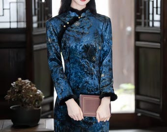 May include: A long, dark blue dress with a floral pattern. The dress has long sleeves with black fur trim at the cuffs and collar. The dress is held closed with small buttons. The person is holding a small, brown book.