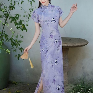 Puede incluir: Vestido chino tradicional lila claro de manga corta, con cuello alto y abertura lateral. El vestido presenta un estampado de pandas y diseños florales. La persona sostiene un pequeño abanico.