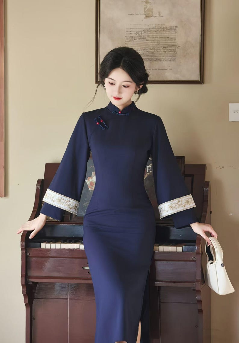 May include: A navy blue, long-sleeved dress with a mandarin collar and a side slit. The dress features embroidered detailing on the sleeves and is worn by a person. A white handbag is held in the right hand. The background includes a piano and a framed picture.