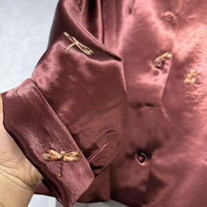 May include: A maroon-colored satin jacket with embroidered dragonfly details on the cuffs. The jacket features small, decorative buttons and subtle floral embroidery near the collar. The fabric has a slight sheen, giving it a luxurious appearance.