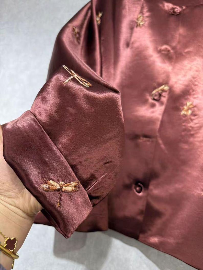 May include: A maroon-colored satin jacket with embroidered dragonfly details on the cuffs. The jacket features small, decorative buttons and subtle floral embroidery near the collar. The fabric has a slight sheen, giving it a luxurious appearance.