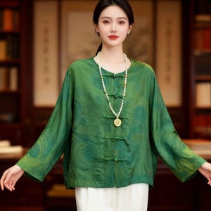 May include: A green long-sleeved blouse with a mandarin collar and frog closures. The blouse has a subtle pattern and is paired with white pants. A long pearl necklace with a gold pendant completes the outfit.
