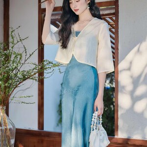 May include: A woman wearing a light blue dress with a matching cream-colored bolero jacket. She is holding a small white beaded purse and a fan. The dress has a floral pattern. The background includes a wooden door frame and a vase with greenery.