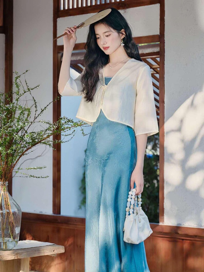 May include: A woman wearing a light blue dress with a matching cream-colored bolero jacket. She is holding a small white beaded purse and a fan. The dress has a floral pattern. The background includes a wooden door frame and a vase with greenery.