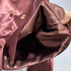 May include: A maroon satin jacket with gold floral embroidery. The jacket features a mandarin collar, decorative buttons, and a smooth, lustrous finish. The sleeve is turned up, revealing a darker, contrasting lining. The garment appears to be part of a formal wear set.