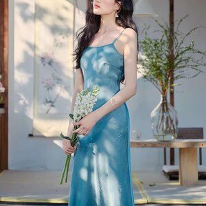May include: A long, light blue dress with spaghetti straps. The dress has a subtle floral pattern and a flowing silhouette. The person is holding a bouquet of white flowers. The background includes a wooden structure and greenery.