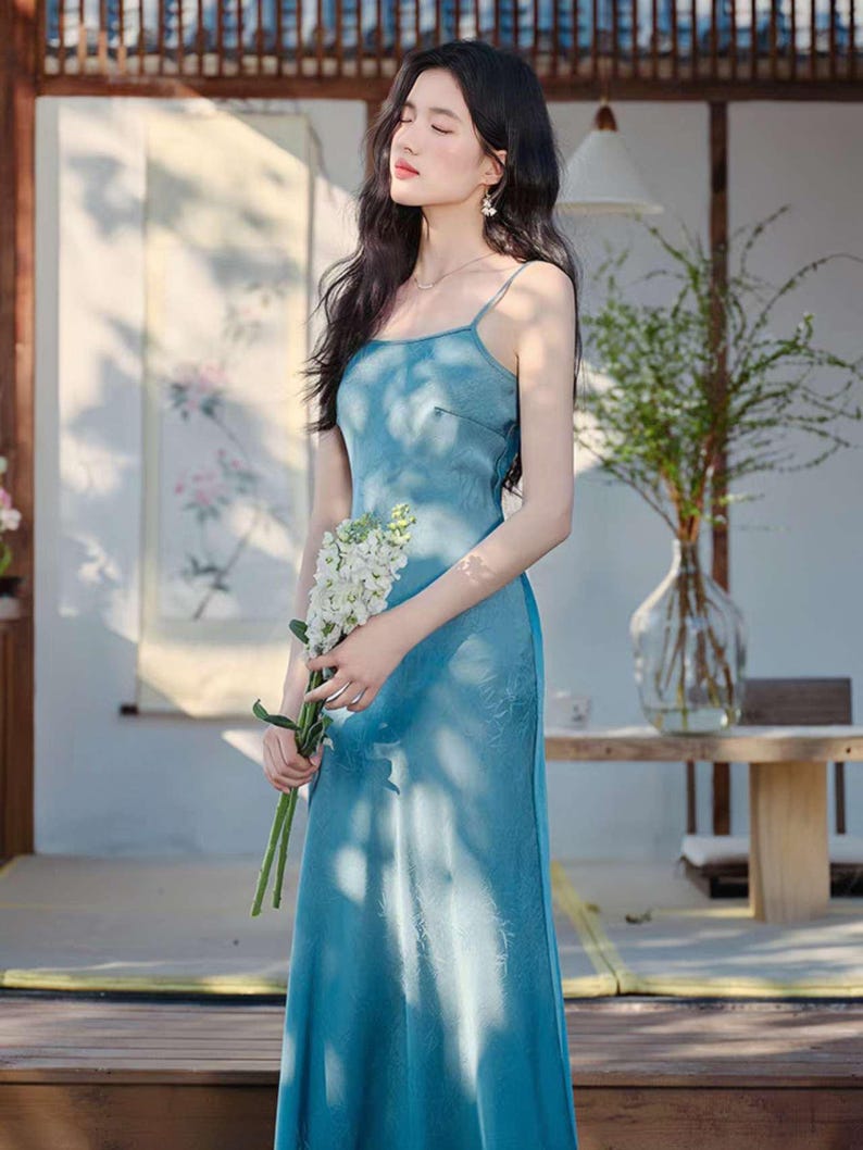 May include: A long, light blue dress with spaghetti straps. The dress has a subtle floral pattern and a flowing silhouette. The person is holding a bouquet of white flowers. The background includes a wooden structure and greenery.