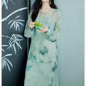 Puede incluir: Un conjunto de dos piezas verde claro con un dise&ntilde;o floral de acuarela. El conjunto incluye una blusa de manga larga con cierres de rana y una falda a juego. La modelo sostiene un plato verde peque&ntilde;o con uvas.