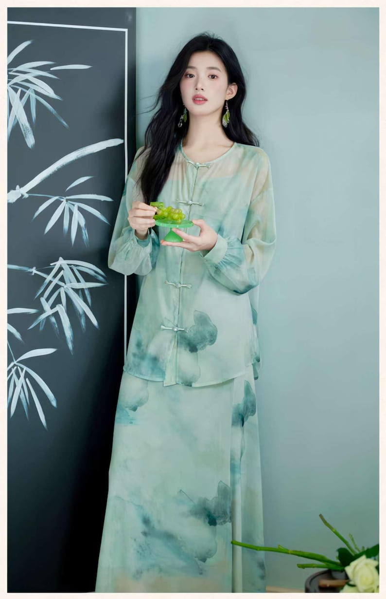 Puede incluir: Un conjunto de dos piezas verde claro con un dise&ntilde;o floral de acuarela. El conjunto incluye una blusa de manga larga con cierres de rana y una falda a juego. La modelo sostiene un plato verde peque&ntilde;o con uvas.