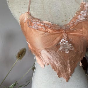 May include: Peach-colored lingerie top with delicate lace and sequin embellishments. The top features a halter neck design and a unique, asymmetrical hemline. The fabric appears to be a combination of silk and sheer mesh, creating a soft, romantic aesthetic.
