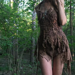 Puede incluir: Un vestido marrón de ganchillo con un dobladillo asimétrico único. El vestido presenta un cuello halter y detalles de encaje intrincados. La prenda está hecha de materiales de aspecto natural, creando una estética rústica y bohemia. El vestido se usa al aire libre en un bosque.