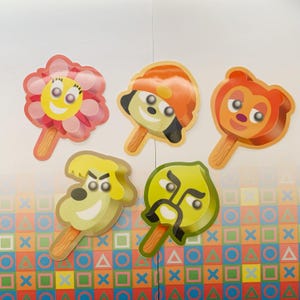 May include: Five colorful cartoon popsicle stickers. The stickers feature various characters, including a flower, a dog, a monkey, and a face with a mustache. Each sticker has a wooden stick and is set against a white background with a colorful pattern.