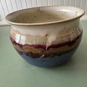 May include: A handcrafted ceramic bowl with a wide, open design. The bowl features a gradient of colors, including a deep blue base, transitioning to brown, red, and a creamy white rim. The pottery has a rustic, organic aesthetic.