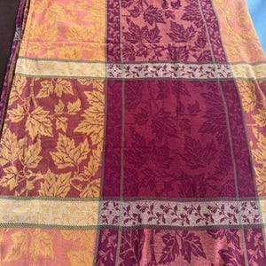 May include: A rectangular fabric tablecloth featuring a woven grid pattern of deep burgundy and golden yellow squares. The design is decorated with an intricate, repeating motif of maple leaves and thin vine-like branches throughout the entire surface.
