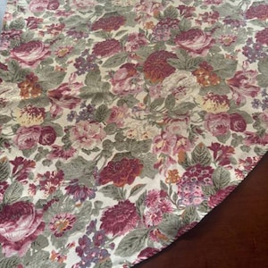 May include: A fabric tablecloth featuring a dense floral pattern with large roses and smaller blossoms in shades of deep burgundy, dusty pink, and muted sage green set against a cream-colored background with visible woven textile texture.