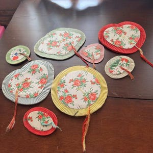 May include: A collection of nine assorted fabric-covered paper fans in various shapes including hearts, circles, and ovals. Each fan features a white fabric center with a red and green floral pattern, surrounded by pleated paper borders in red, yellow, green, and blue, with attached decorative tassels.