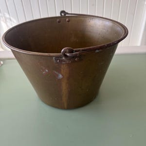 May include: A vintage brass bucket with a conical shape and a sturdy handle. The bucket has a tarnished patina, showing signs of age and use. The handle is attached with rivets, adding to its rustic charm. This bucket is a decorative item.